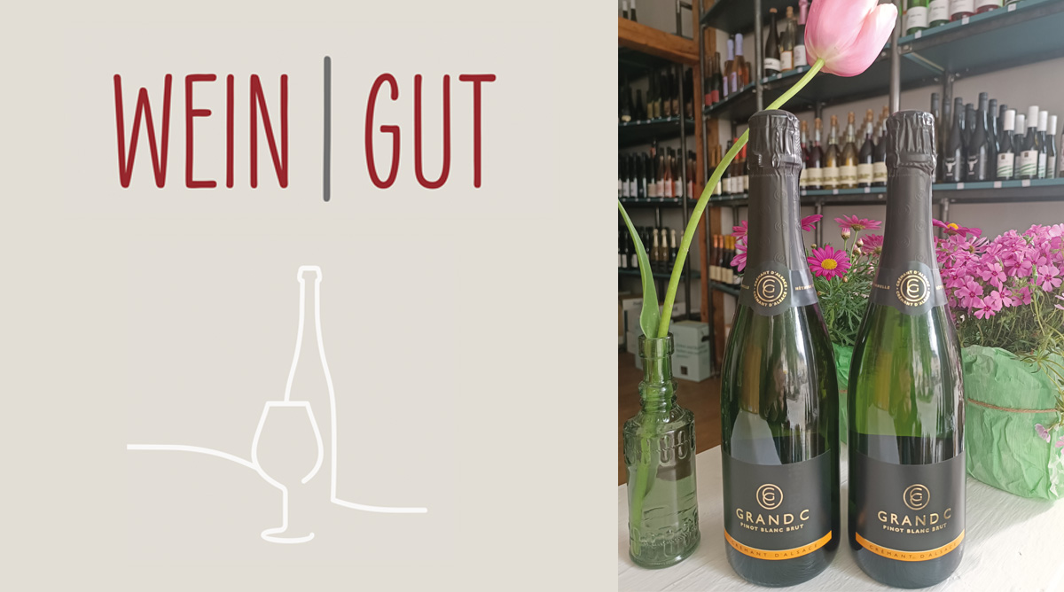 Zwei Flaschen Grand C Pinot Blanc Brut stehen auf einem Tisch, umgeben von Blumen und einem Glas. Der Hintergrund zeigt Weinregale.