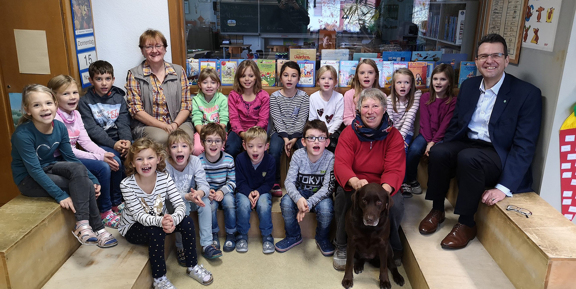 Gruppenfoto von Kindern, Lehrern und einem Hund in einem Klassenzimmer, alle lächelnd und fröhlich.