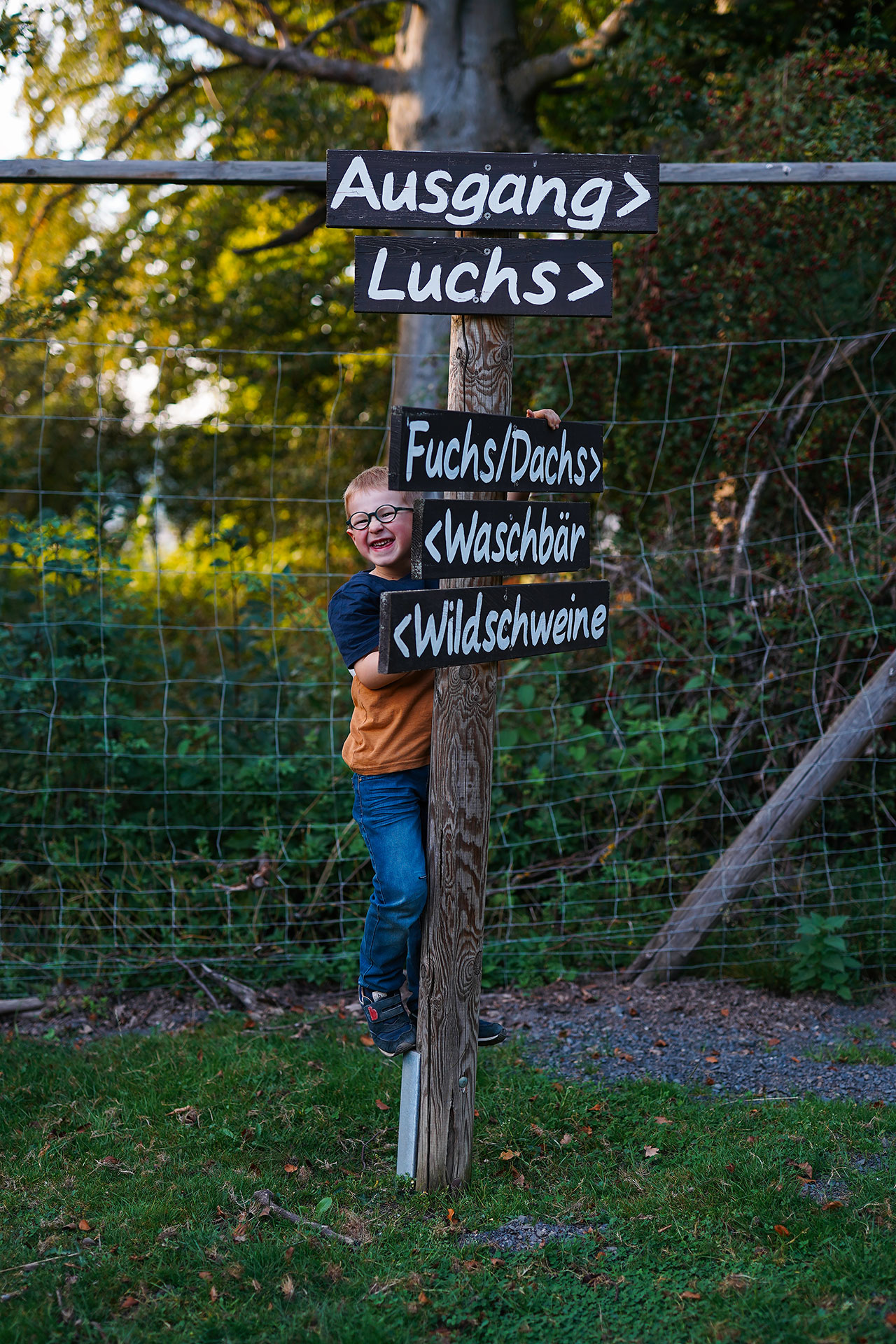 Lachender Junge, der an einem Holzpfahl mit Schildern zu Tieren und einem Ausgang lehnt, umgeben von Natur.