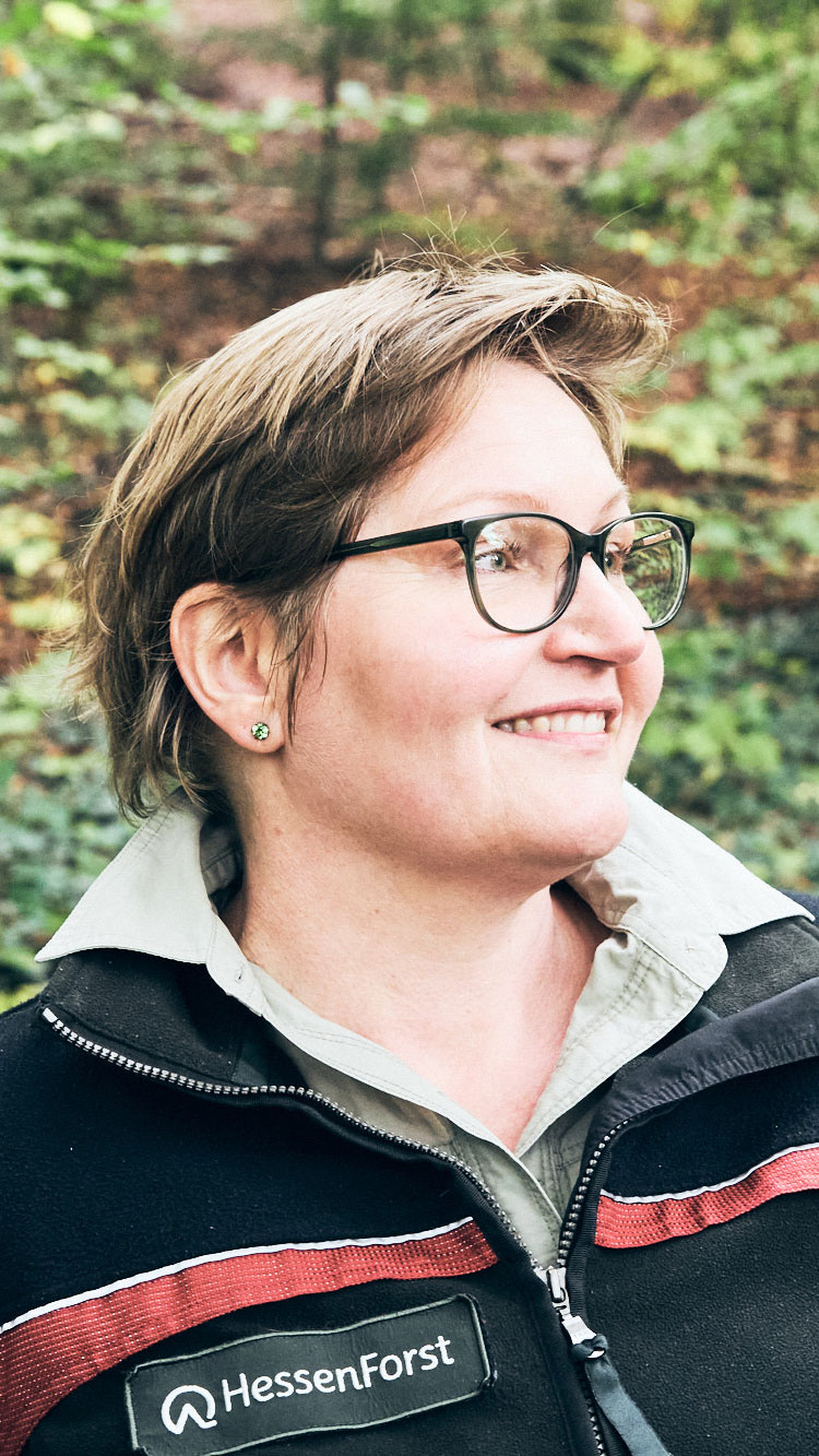 Porträt einer lächelnden Frau mit kurzen Haaren, Brille und Arbeitskleidung in einem Waldumfeld.