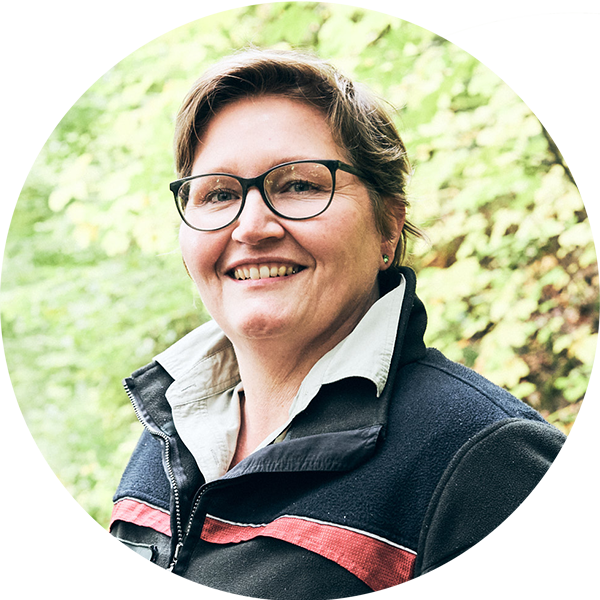 Porträt einer lächelnden Frau mit Brille und Fleecejacke, umgeben von grüner Natur.