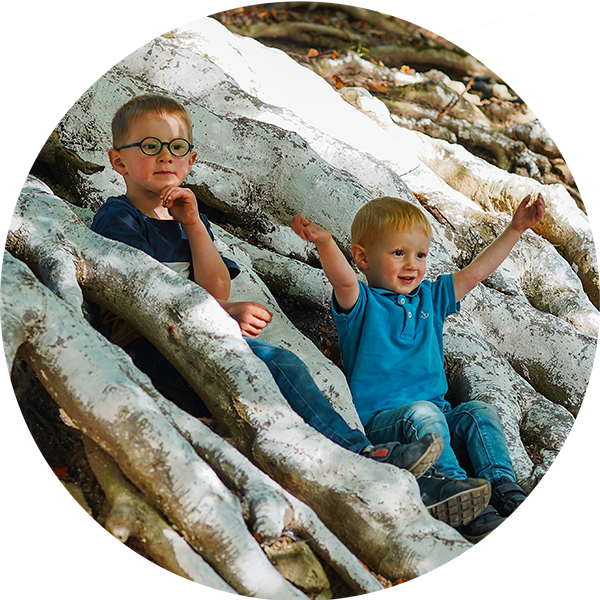 Zwei Kinder sitzen auf großen Wurzeln, der ältere schaut nachdenklich, der jüngere freut sich und hebt die Arme.