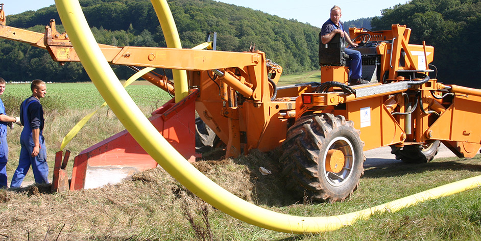 Arbeiter nutzen eine orangefarbene Maschine zum Graben, während ein grünes Feld und Hügel im Hintergrund sichtbar sind.