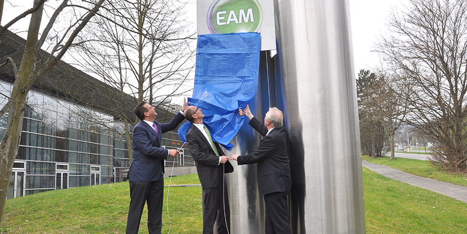 Drei Männer enthüllen gemeinsam ein Schild mit dem Schriftzug "EAM"" an einem hohen