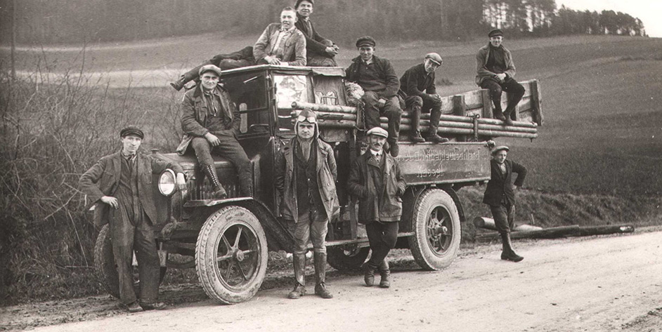 Schwarzweißfoto von acht Männern auf einem alten Lastwagen, umgeben von ländlicher Umgebung.