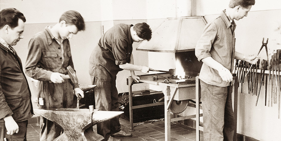 Vier Handwerker in einer Schmiede, die Metall bearbeiten und Werkzeuge nutzen, um handwerkliche Arbeiten auszuführen.