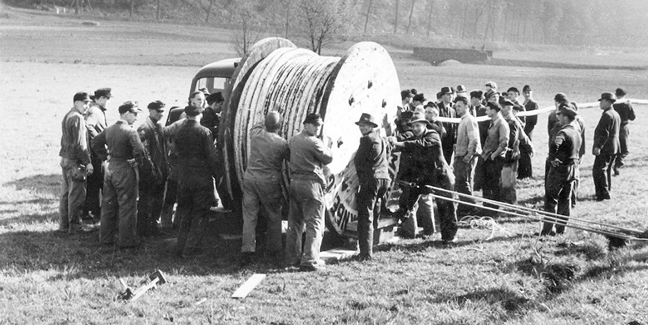 Gruppierung von Menschen um eine große Kabeltrommel auf einer Wiese, in Schwarz-Weiß.