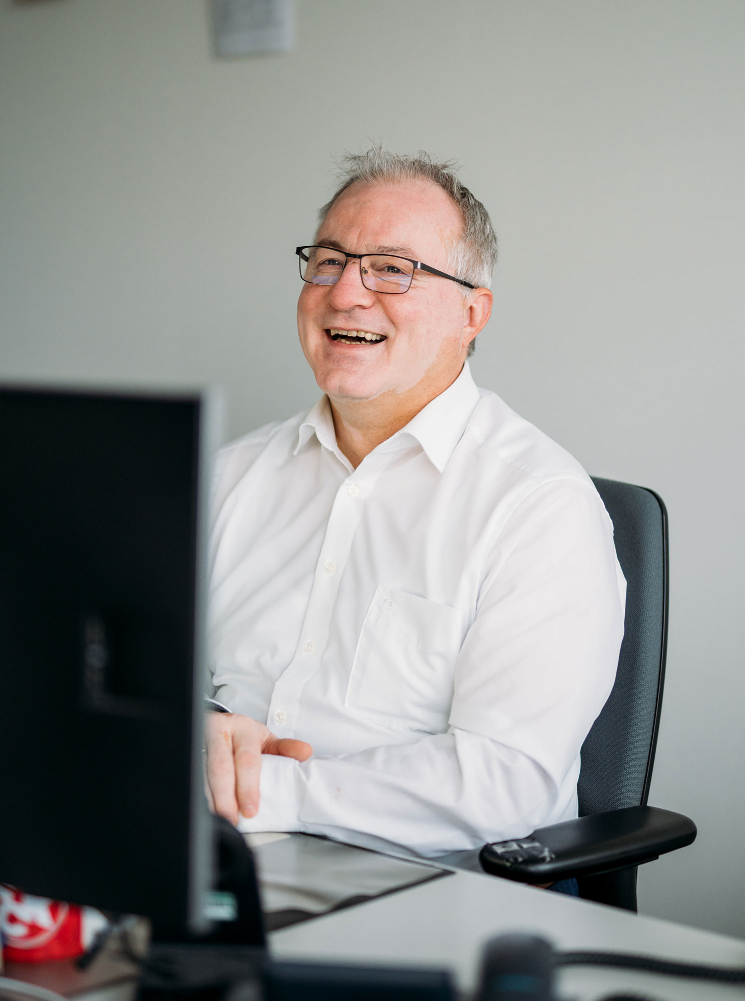 Lächelnder Mann in weißem Hemd sitzt am Schreibtisch vor einem Computer, mit einer entspannten Haltung.