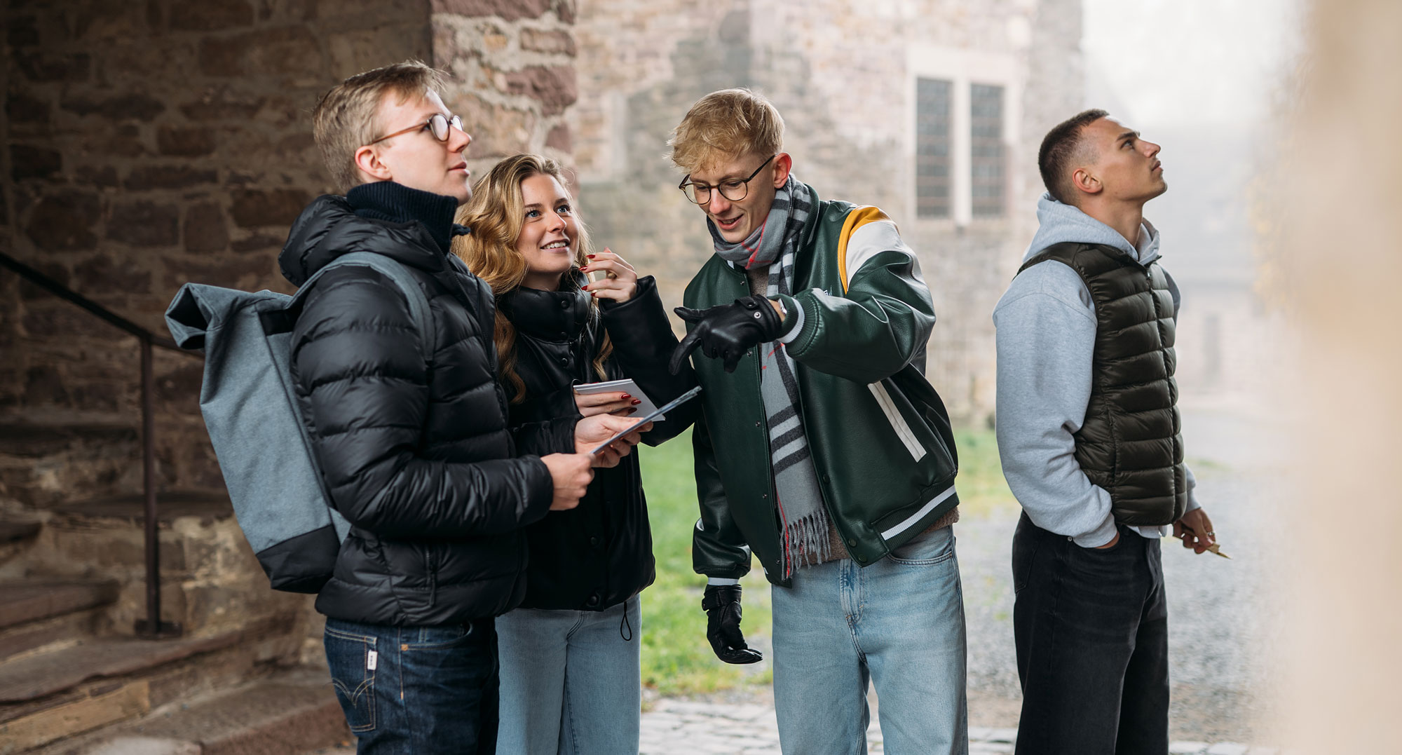 Vier junge Erwachsene stehen in einem Innenhof und unterhalten sich, während einer ein Tablet hält und auf etwas zeigt.