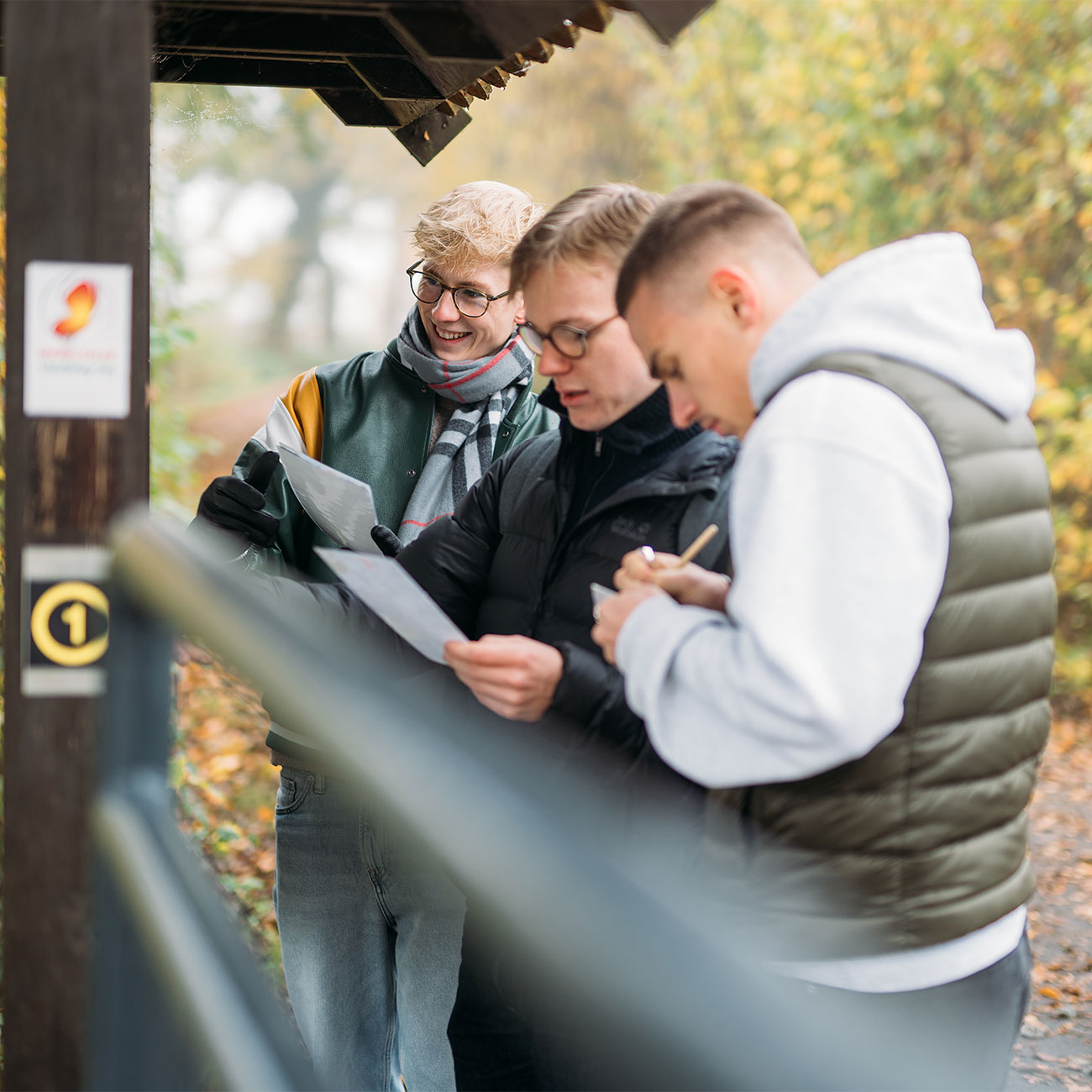 Drei junge Männer stehen unter einem Vordach, schauen auf Dokumente und sprechen miteinander bei herbstlichem Wetter.