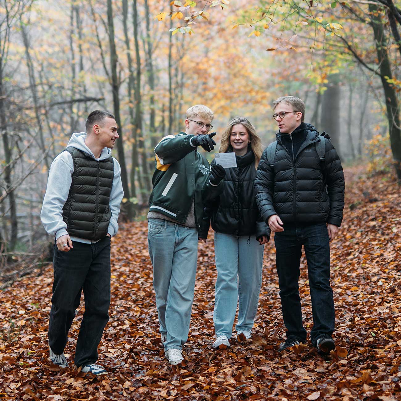 Vier junge Menschen lachen und interagieren auf einem mit Herbstlaub bedeckten Weg im Wald.