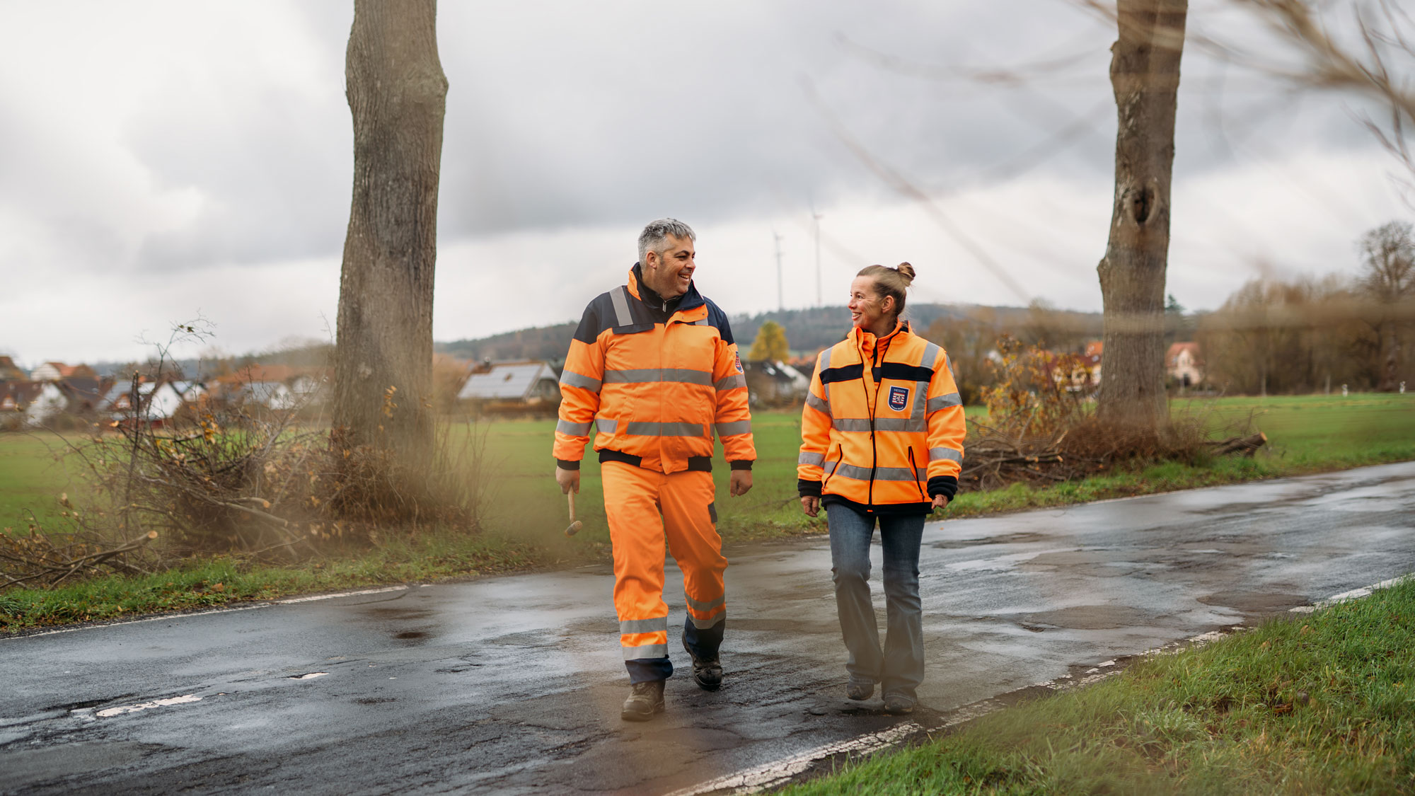 Zwei Personen in orangefarbener Arbeitskleidung gehen auf einer Landstraße entlang, umgeben von einer ländlichen Landschaft.