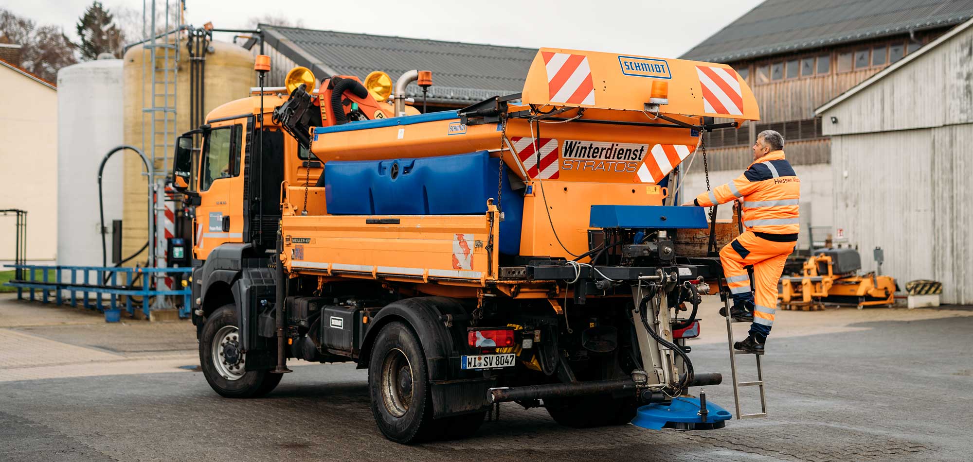 Ein orangefarbener Winterdienst-Lkw mit einem Arbeiter, der eine Treppe hinaufsteigt, um den Gerätebereich zu erreichen.