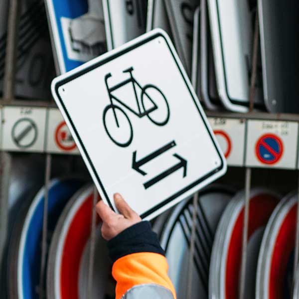 Person hält ein Verkehrsschild mit einem Fahrrad und einem Pfeil nach links in einer Reihe von Schildern.