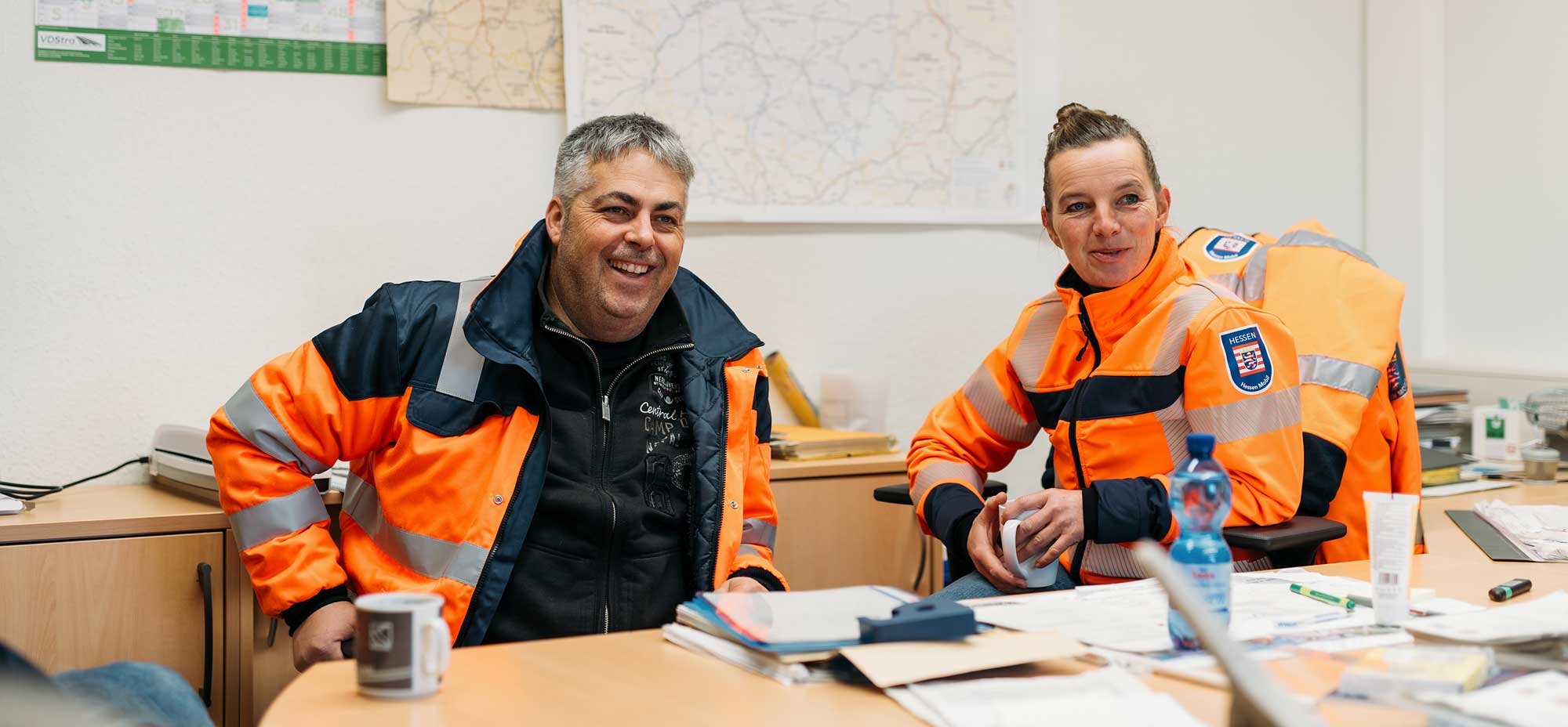 Zwei Personen in orangefarbener Arbeitskleidung sitzen an einem Tisch mit Unterlagen und Tassen vor ihnen. Sie lächeln.
