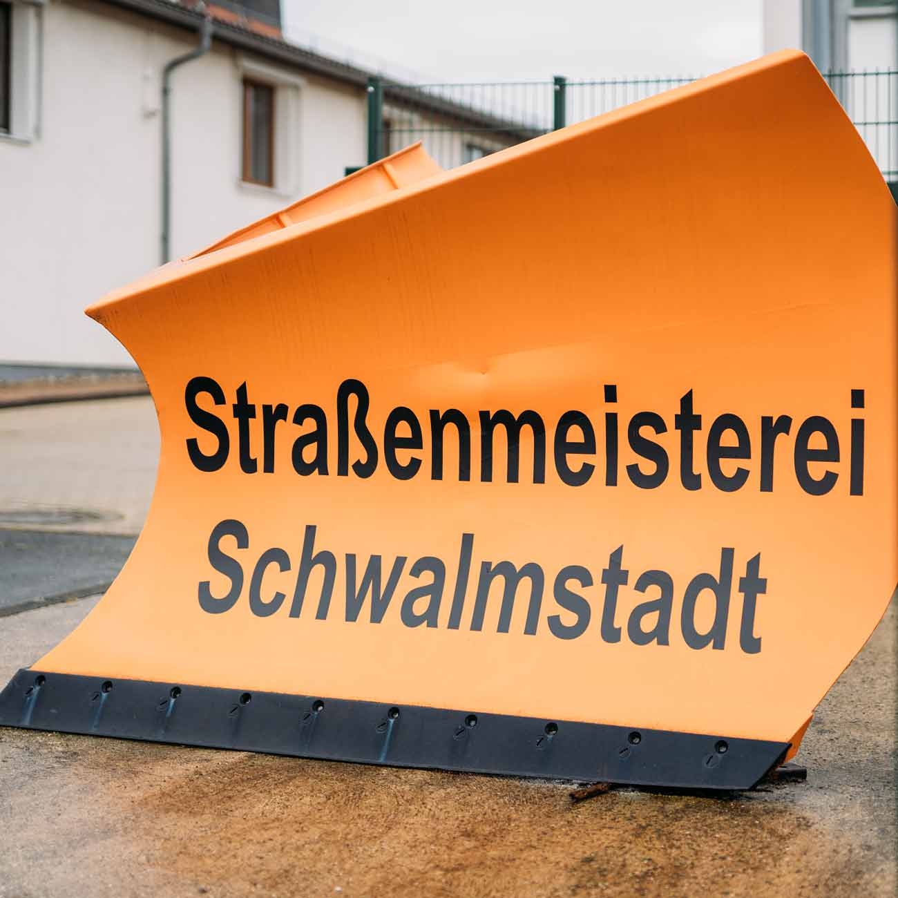 Orangefarbener Schild mit dem Text "Straßenmeisterei Schwalmstadt"" auf einem gepflasterten Untergrund."