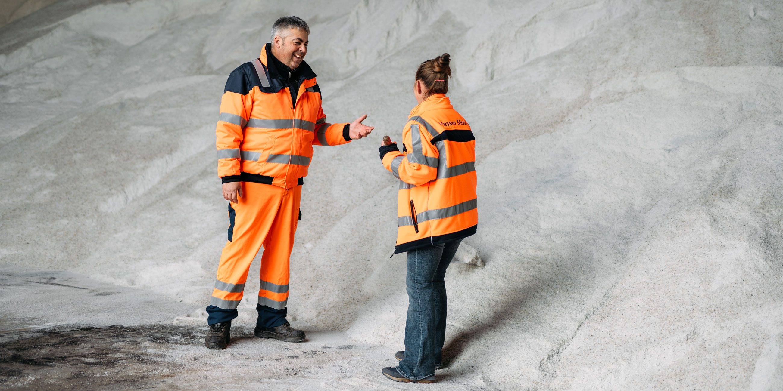 Zwei Sicherheitskräfte in orangefarbenen Overalls unterhalten sich vor einem großen Salzvorrat.