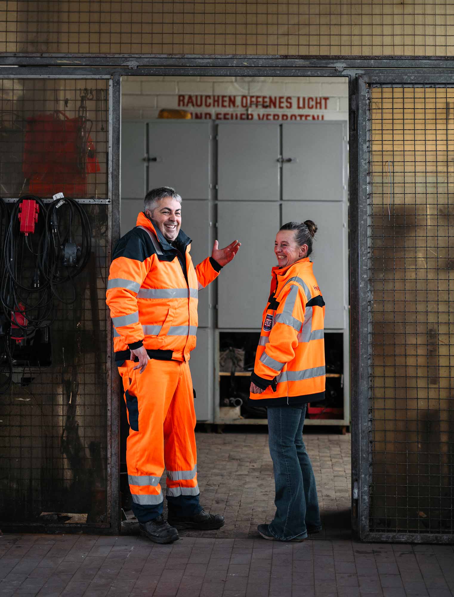 Zwei Personen in leuchtend orangen Arbeitsjacken stehen in einem Eingang und lachen miteinander. Im Hintergrund ist eine geschlossene Tür.