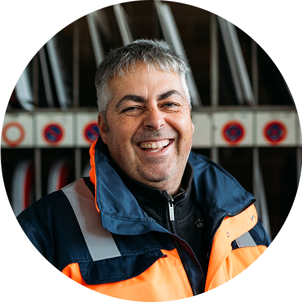 Lächelnder Mann in orangefarbener Arbeitsjacke vor einem Regal mit Schildern.