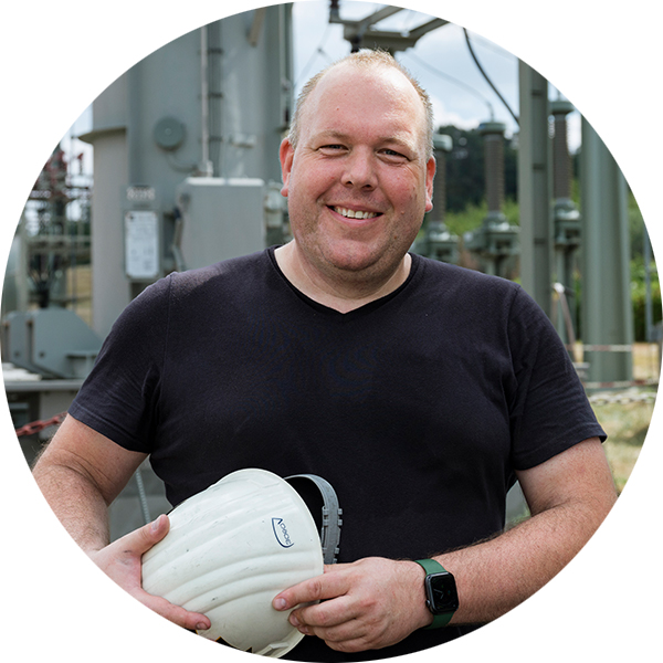 Mann mit Helm in der Hand, lächelt vor einer technischen Kulisse im Freien.