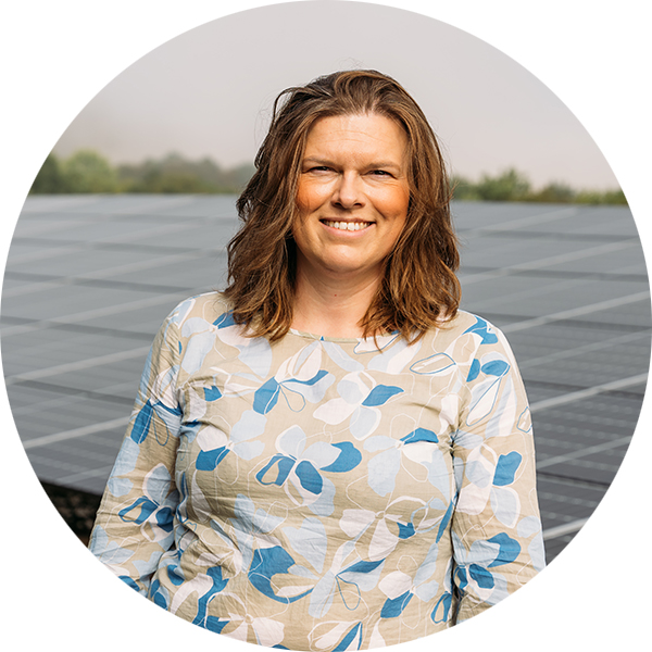 Frau mit braunen Haaren, lächelnd vor Solarpanelen, trägt ein blumiges Oberteil in Blau- und Beigetönen.