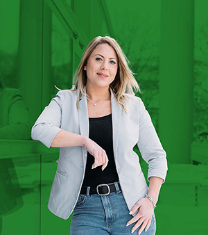 Frau mit blonden Haaren in grauem Blazer, lächelnd, vor einer grünen Wand stehend.