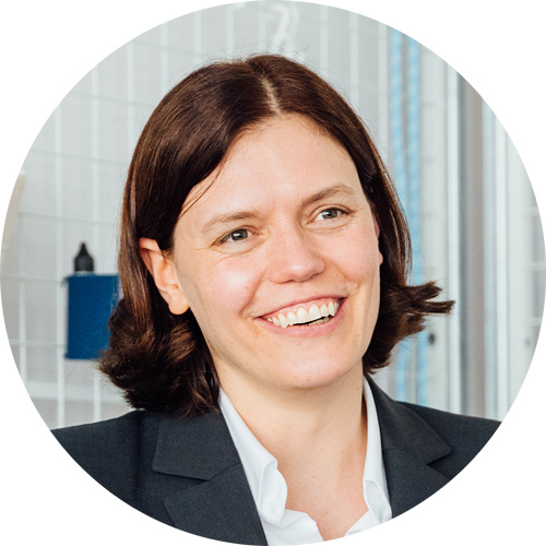 Lächelnde Frau mit dunklen Haaren, die im Büroportrait in einem schwarzen Blazer abgebildet ist.