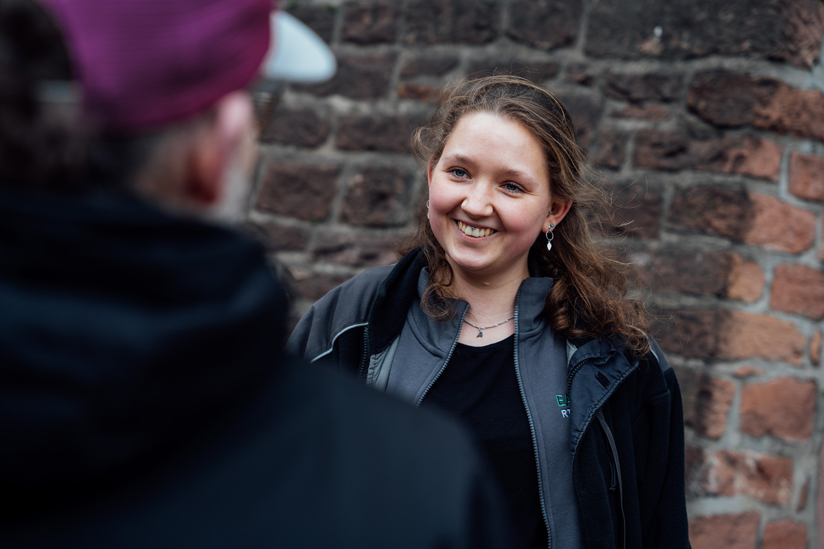 Lächelnde Frau mit lockigem Haar in einem grauen Jackett, im Gespräch vor einer steinernen Wand.