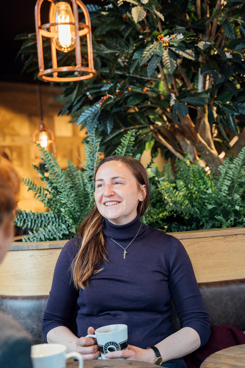 Lächelnde Frau mit langen Haaren sitzt in einem Café, hält einen Kaffeebecher, umgeben von Pflanzen und sanfter Beleuchtung.