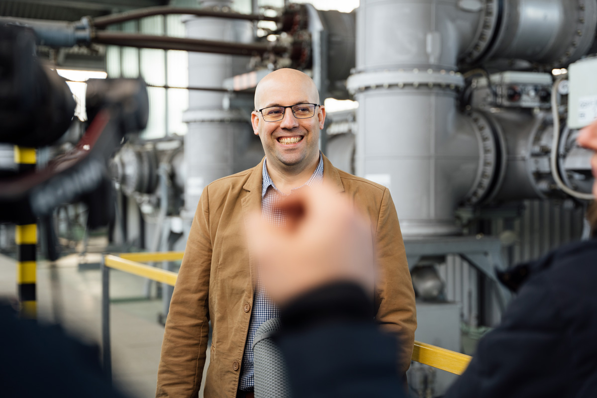 Lächelnder Mann in einem Industriebereich, umgeben von großen Maschinen und Rohren.