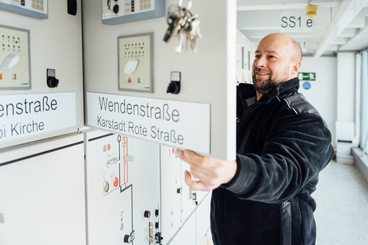 Mann in schwarzer Jacke, der ein Bedienfeld mit beschrifteten Schaltern in einem modernen technischen Raum bedient.