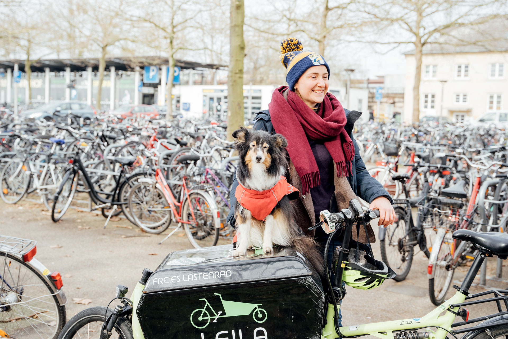 Frau mit Hund auf einem Lastenrad, umgeben von einer Fahrradparkplatze in einer städtischen Umgebung.