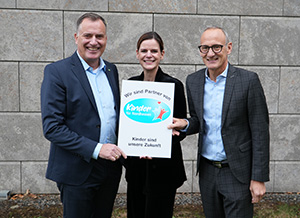 Der EAM-Geschäftsführer Olaf Kieser, die Leiterin des Bereichs Sponsoring der EAM und der Chef der HNA stehen vor einer grauen Steinwand und halten gemeinsam ein Schild. Auf dem Schild steht: ‚Wir sind Partner von Kinder für Nordhessen – Kinder sind unsere Zukunft‘, begleitet von einem Logo mit einer stilisierten Figur und blauen sowie roten Akzenten.