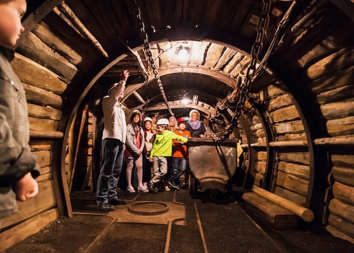 Gruppe von Menschen in einem Holzstollen, umgeben von Bergbaumaschinen und dimmem Licht.