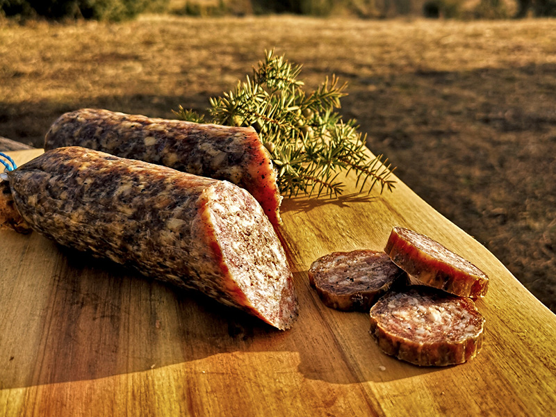 Hausgemachte Wurst auf einem Holzbrett, umgeben von frischem Grün, mit ein paar Scheiben daneben.