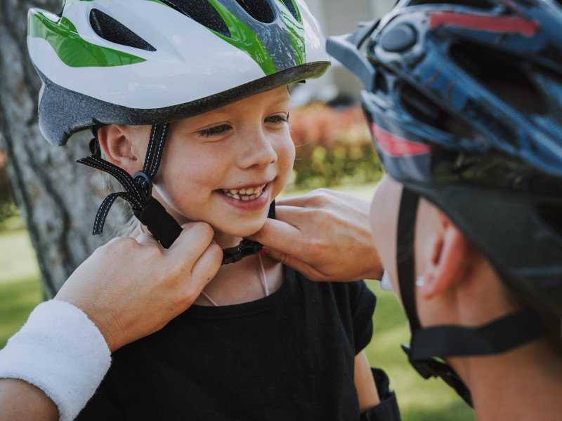Ein Kind lächelt, während eine Person ihm einen Fahrradhelm anlegt, im Freien vor verschwommenem Hintergrund.