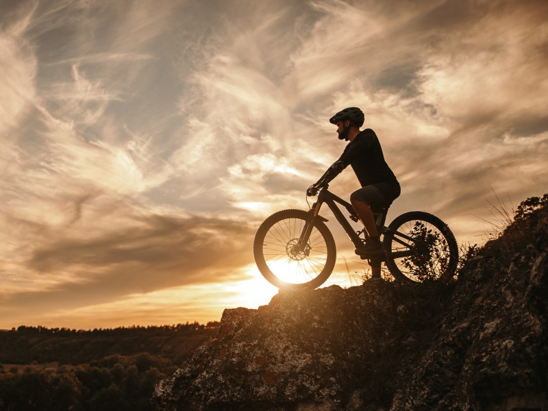 Ein Mountainbiker steht auf einem Felsen, mit Sonnenuntergang und Wolken im Hintergrund.