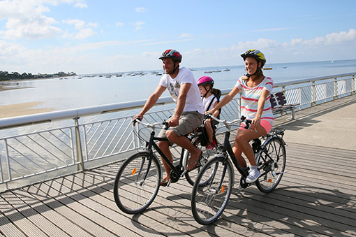 Familie fährt auf Fahrrädern entlang einer Uferpromenade, sonniger Tag, Wasser im Hintergrund.