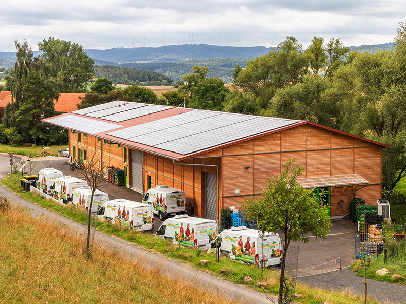 Holzgebäude mit Solarpanelen und Lieferwagen auf einem Weg, umgeben von Bäumen und Feldern.
