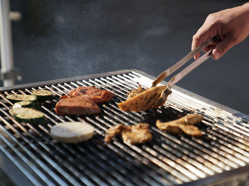 Hand eines Grillenden mit Zange, die mariniertes Fleisch und Gemüse auf einem Grill wendet, Rauch steigt auf.