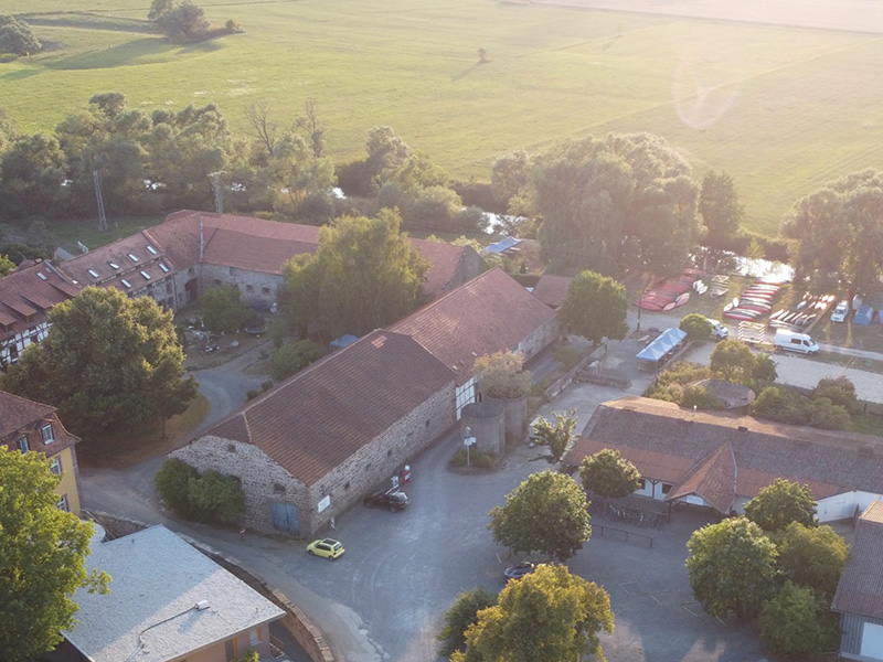 Luftaufnahme eines landwirtschaftlichen Gebäudekomplexes umgeben von grünen Feldern und Bäumen, mit Fahrzeugen und Booten im Hintergrund.