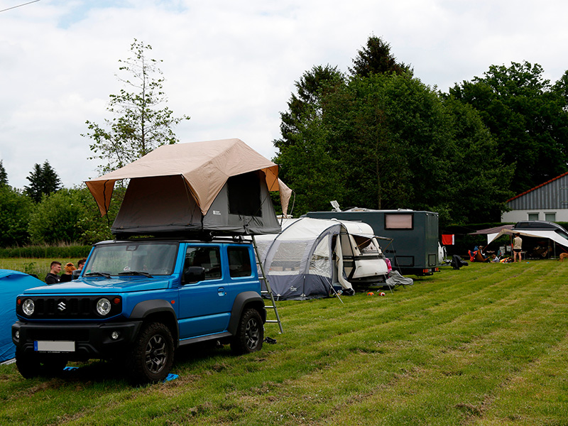 Blaues Auto mit Klappdach, umgeben von Zelten auf einer grünen Wiese in einer entspannenden Campingumgebung.