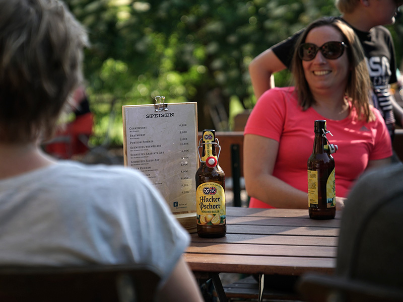 Zwei Frauen am Tisch mit Bierflaschen und einer Speisekarte im Freien, umgeben von Pflanzen.