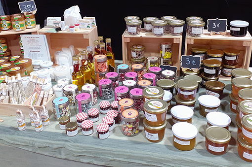 Verschiedene Gläser mit Marmelade, Honig und Ölen auf einem Tisch, umgeben von Holzregalen und Preisschildern.