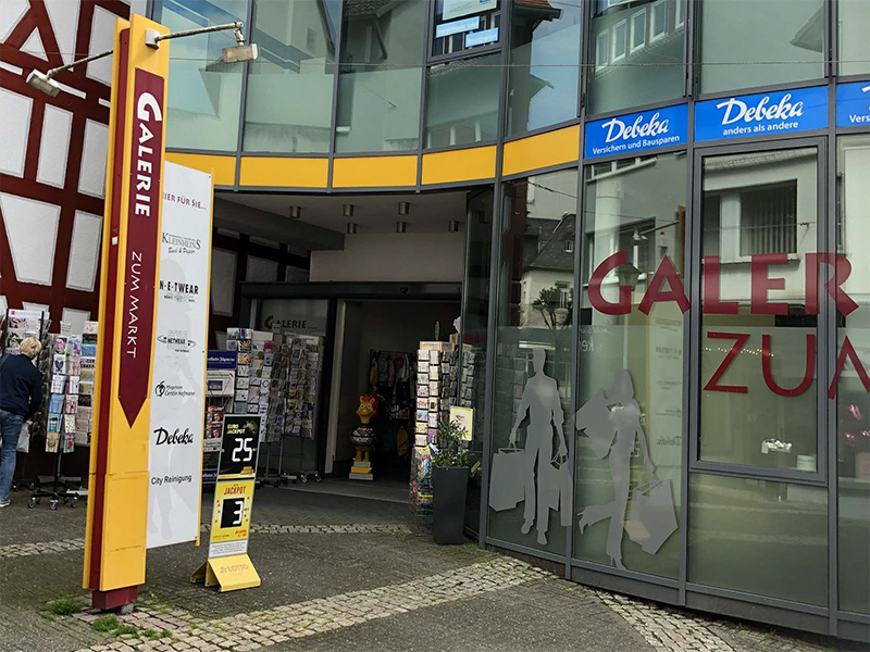 Eingang zur Galerie zum Markt mit Schildern und Auslagen in einem modernen Gebäude. Menschen stehen vor dem Eingang.