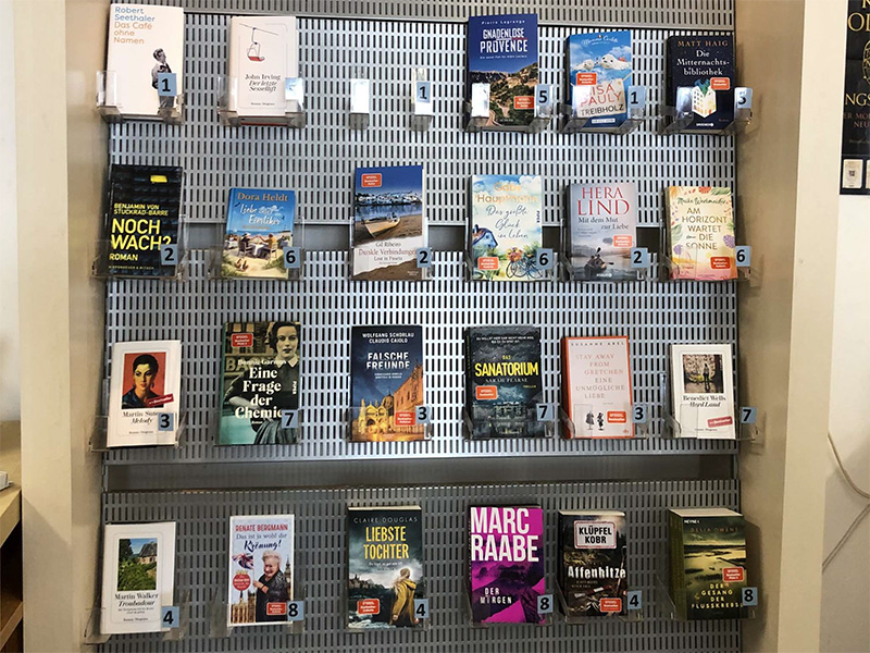 Regal mit acht Bücherständern, die verschiedene Buchcover zeigen, sowie Preisschilder in einer Bibliothek.