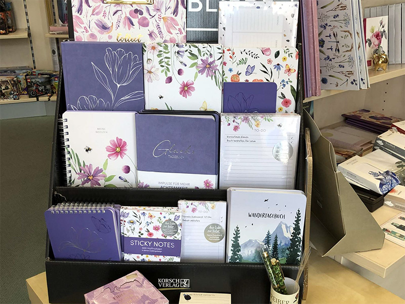 Regal mit verschiedenen Notizbüchern und Schreibwaren in lila und blumigem Design.