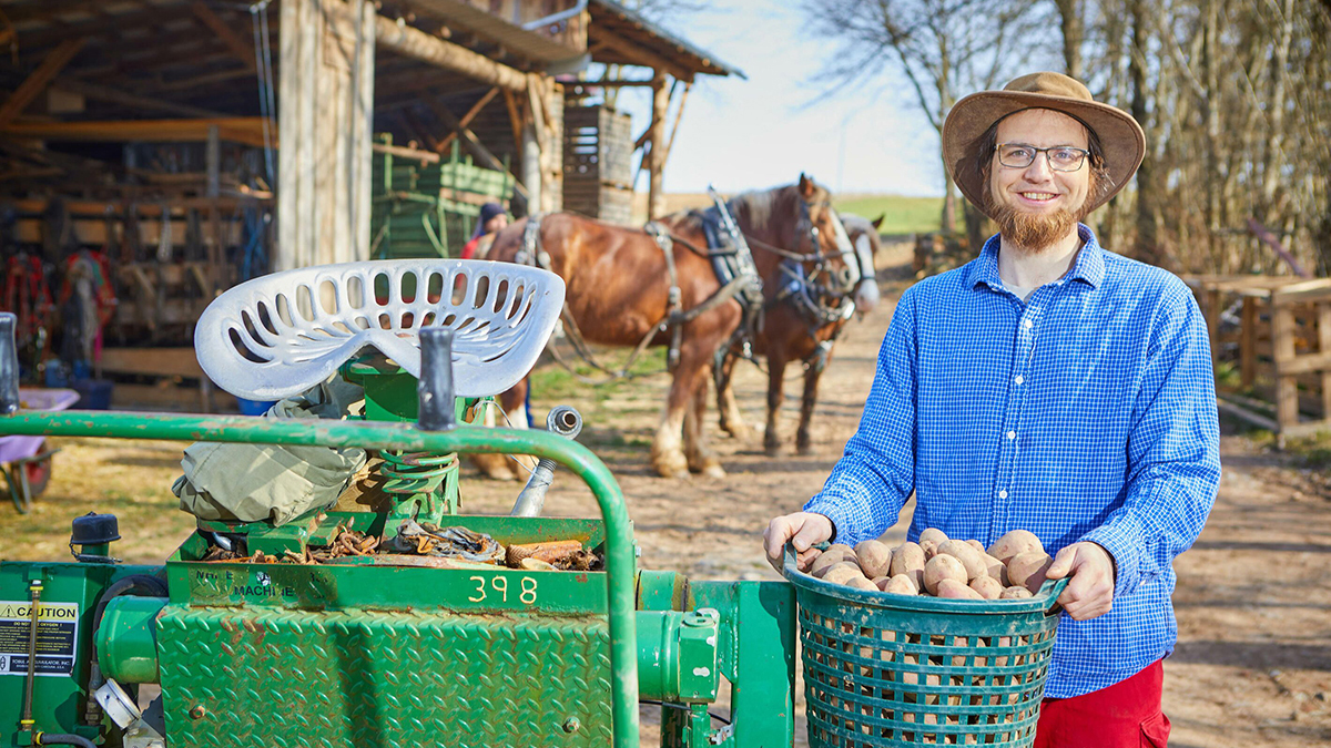 Ein lächelnder Mann mit Brille und Hut steht vor einer Kartoffelmaschine und hält einen Korb mit Kartoffeln. Im Hintergrund sind Pferde.