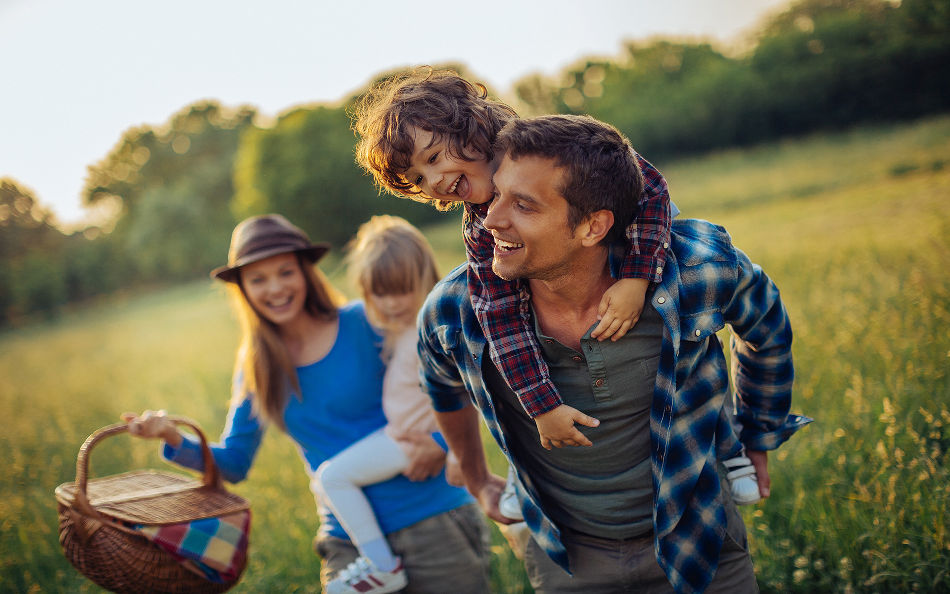 Familie mit zwei Kindern in einem Feld, der Vater trägt den Sohn auf dem Rücken, die Mutter hält einen Picknickkorb.