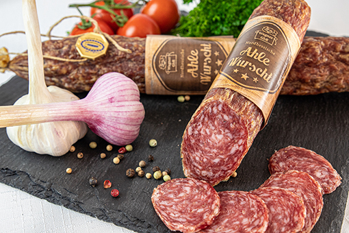 Scheiben einer Wurst auf einem schwarzen Steinbrett, umgeben von Knoblauch, Pfefferkörnern und Tomaten.